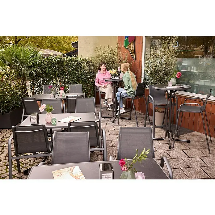 MFG Garten-Stehtisch rund - Ø x H: 70 x 110 cm, Stahl, Mecalit, Rund, Schiefer - Einladende Außenansicht eines Cafés mit grauen Tischen und Stühlen, dekoriert mit Blumen, wo sich zwei Personen unterhalten.