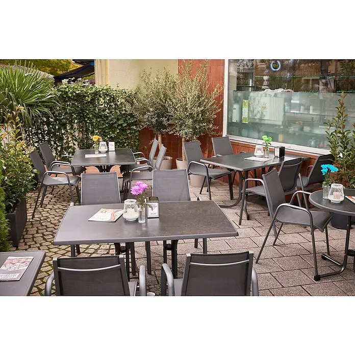 MFG Gartentisch Boulevard - L x B: 132 x 90 cm, Stahl, Mecalit, Schiefer, Klappbar - Außenaufnahme eines Restaurants mit Tischen und Stühlen, die zum Verweilen einladen, umgeben von Pflanzen, die eine angenehme Atmosphäre schaffen.