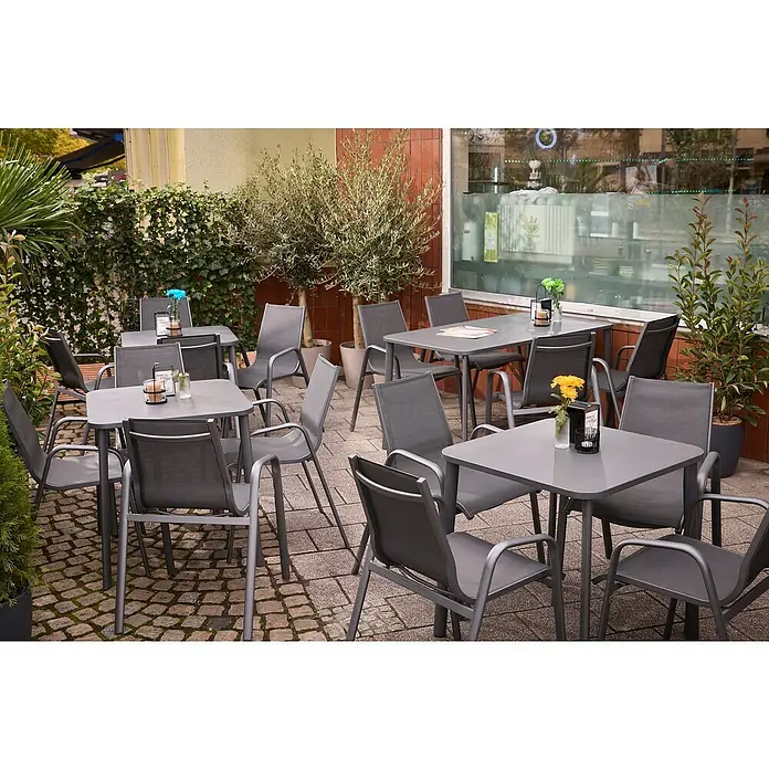 Sunfun Gastro Gartentisch - L x B x H: 74,5 x 75 x 74,5 cm, Stahl, Night-Grey, Stapelbar - Außenbereich eines Cafés mit grauen Tischen und Stühlen, umgeben von Pflanzen für eine entspannte Atmosphäre.