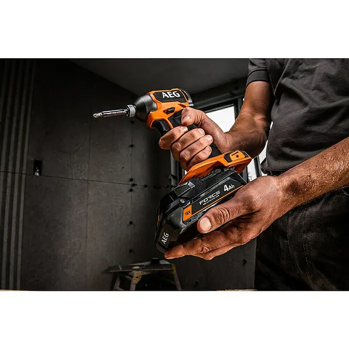 AEG Pro 18V Akku FORCE PROLITHIUM-ION™ - 4 Ah, Li-Ionen - Nahaufnahme eines AEG Akku-Schlagschraubers mit orangefarbenen Akzenten, gehalten von einer Hand vor einem Bauhintergrund.