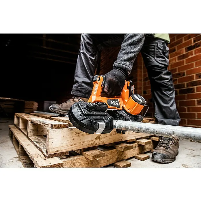 AEG Pro 18V Akku-Bandsäge BNS 18SBL-0 - 18 V, Ohne Akku - Nahaufnahme einer AEG Akku-Bandsäge beim Schneiden eines Metallrohrs, bedient von einer Person in Arbeitskleidung auf einer Holzpalette.