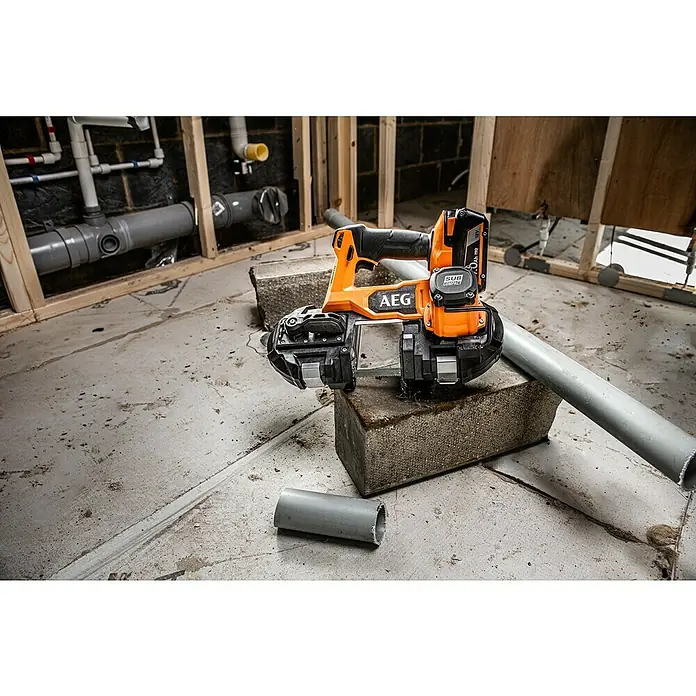 AEG Pro 18V Akku-Bandsäge BNS 18SBL-0 - 18 V, Ohne Akku - Nahaufnahme einer AEG-Bandsäge auf Ziegelsteinen, die ein graues Rohr schneidet, im Hintergrund Rohrarbeiten.