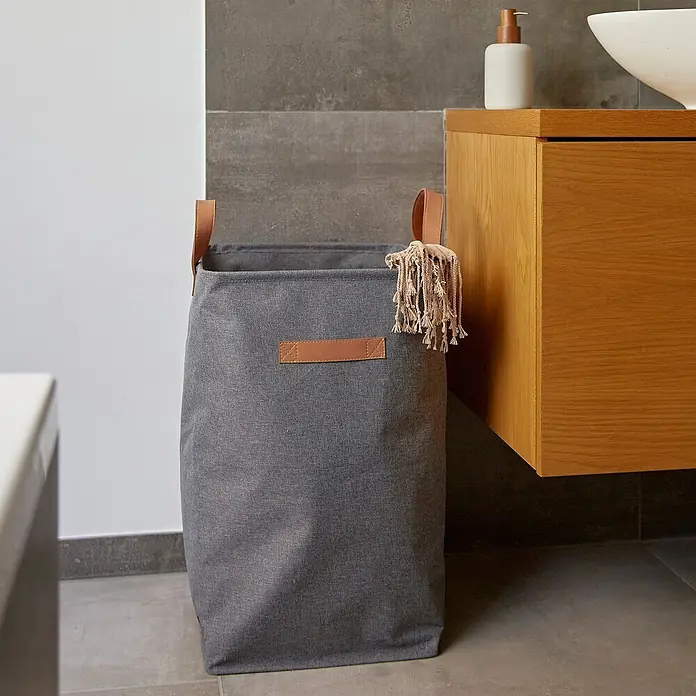 Store It Premium Wäschekorb - L x B x H: 35 x 35 x 58 cm, Grau - Ein grauer Wäschekorb aus Stoff mit braunen Ledergriffen steht im Badezimmer. Eine Quaste hängt am Korb.