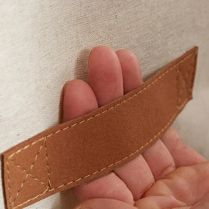 Store It Premium Wäschekorb - L x B x H: 35 x 35 x 58 cm, Beige - Nahaufnahme eines hellbraunen Wildledergriffs mit goldenen Nähten, der an einem beigen Stoff befestigt ist und von einer Hand gehalten wird.