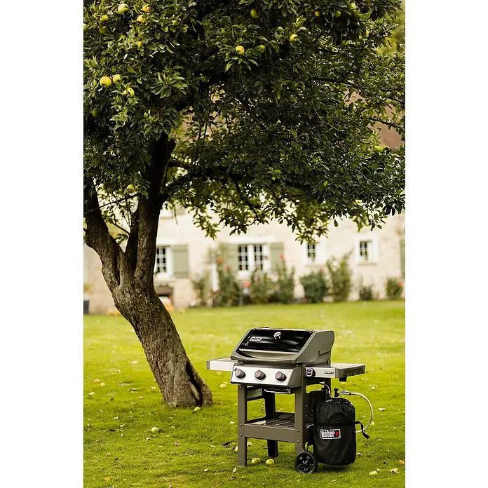 Weber Barbacoa de gas Spirit II E-310 + 7 accesorios - Superficie de la parrilla (An x Pr): 60 x 46 cm, 8,7 kW, Número de quemadores: 3 - Barbacoa de gas Weber Spirit II E-310 debajo de un manzano. La barbacoa es gris, tiene tres quemadores y una bolsa de gas negra.