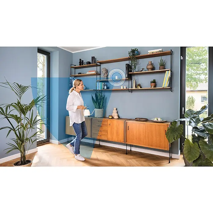 Bosch Smart Home Bewegungsmelder II [+M] Matter - weiß - Eine Frau gießt Pflanzen in einem Wohnzimmer mit einem smarten Home-Sensor an der Wand, der die Raumumgebung überwacht.