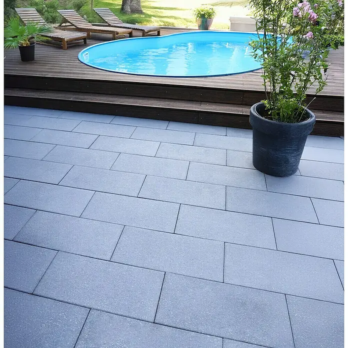 EHL Terrassenplatte Ansina - 60 x 40 x 4 cm, Anthrazit, Beton - Ein ebenerdiger Außenbereich mit grauen Pflastersteinen, der zu einem Poolbereich mit Holzdeck und Liegestühlen führt. Ein großer Topf mit blühenden Pflanzen steht.