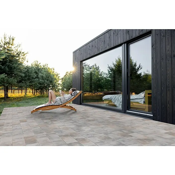 EHL Terrassenplatte Protect Light - 60 x 30 x 4 cm, Muschelkalk, Beton - Frau entspannt auf einer Outdoor-Liege auf einer Steinterrasse neben einem modernen Haus mit großen Fenstern und genießt die Aussicht.