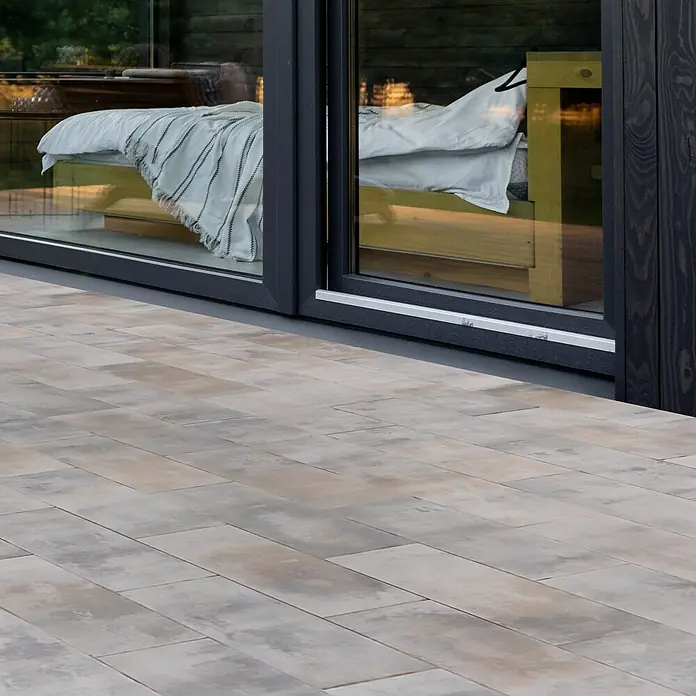 EHL Terrassenplatte Protect Light - 60 x 30 x 4 cm, Muschelkalk, Beton - Die Aufnahme zeigt Pflastersteine im Vordergrund und ein Fenster mit Blick auf ein Bett im Hintergrund. Die Steine sind in verschiedenen Grautönen gehalten.