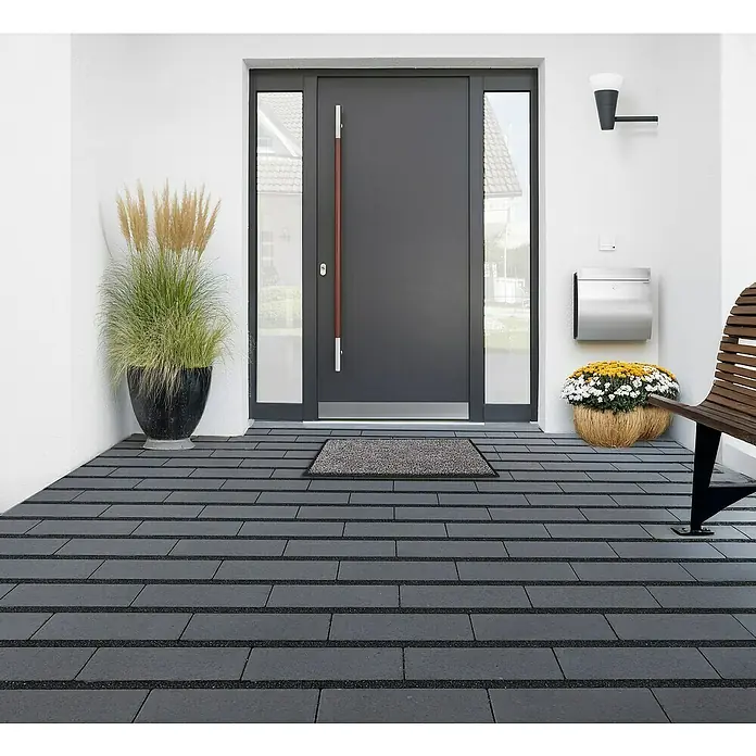 EHL Pflasterstein Cortona Drän - 30 x 15 x 8 cm, Anthrazit, Beton - Hauseingang mit grauer Tür, Pflanzkübeln und Steinboden. Die moderne Haustür lädt ein und die Bepflanzung sorgt für eine freundliche Atmosphäre.