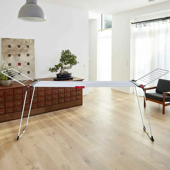 Vileda Flügel-Wäscheständer Infinity Flex - Länge Wäscheleine: 30 m, L x B x H: 304 x 56 x 128 cm, Weiß - Ein Vileda Wäscheständer in einem hellen Raum. Der Wäscheständer ist weiß und rot. Im Hintergrund sieht man ein Sideboard, einen Sessel und Vorhänge.