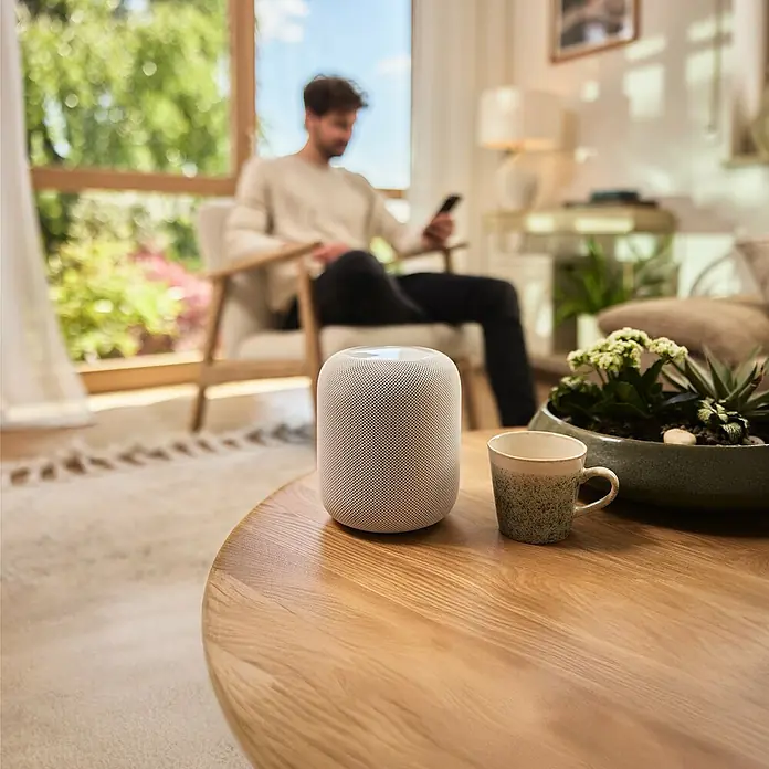 Gardena Smart system Bewässerungsautomat Dual Water Control - Betriebsdruck: 0,5 bar - 12 bar, Bewässerungsdauer: 1 min - 10 h - Ein smarter Lautsprecher steht auf einem Holztisch, im Hintergrund ein Mann mit einem Smartphone.
