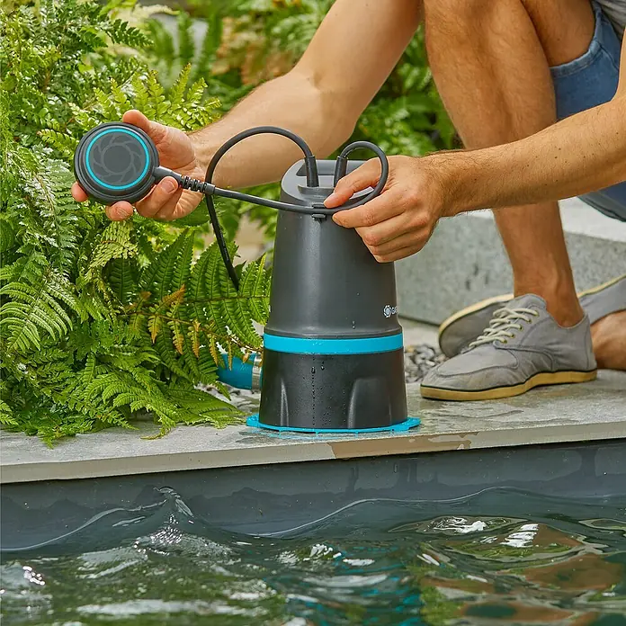 Gardena Schmutzwasserpumpe 20000 Basic Hochwasser-Set - 550 W, Max. Fördermenge: 20.000 l/h, Eintauchtiefe: 7 m - Ein Mann bedient eine graue und blaue Gardena Teichpumpe am Rande eines Teiches, mit einem Sensor in der Hand.