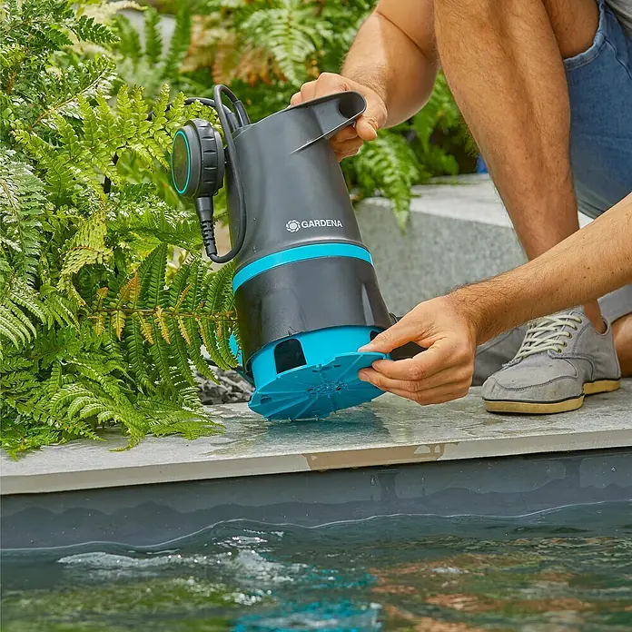 Gardena Schmutzwasserpumpe 20000 Basic Hochwasser-Set - 550 W, Max. Fördermenge: 20.000 l/h, Eintauchtiefe: 7 m - Eine GARDENA Schmutzwasserpumpe wird in einen Teich abgesenkt, um das Wasser abzupumpen. Die Pumpe ist schwarz und blaugrün.