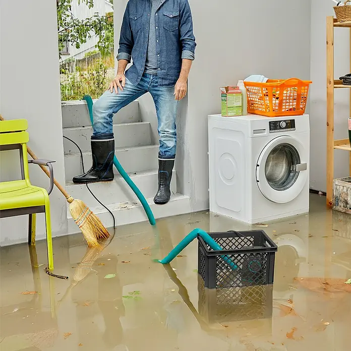Gardena Schmutzwasserpumpe 20000 Basic Hochwasser-Set - 550 W, Max. Fördermenge: 20.000 l/h, Eintauchtiefe: 7 m - Ein Mann in Gummistiefeln steht in einem überfluteten Raum mit einer Pumpe. Die Waschmaschine steht auch im Wasser.