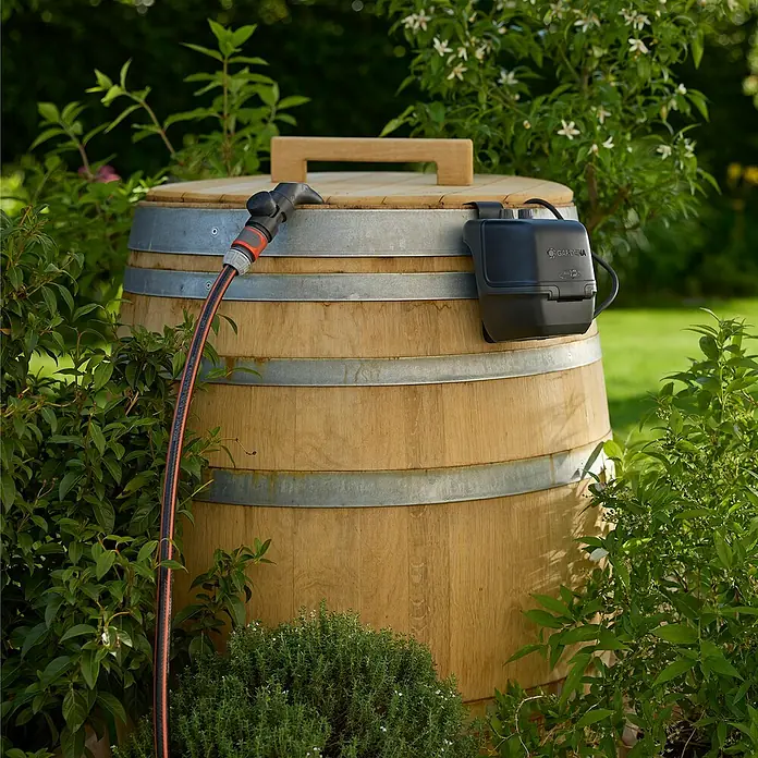Gardena Power for All 18V Akku-Regenfasspumpe 2500 ComfortAutomatic - Ohne Akku, Max. Fördermenge: 2.400 l/h - Nahaufnahme eines Holzfasses mit Bewässerungssystem und Schlauch im Garten.