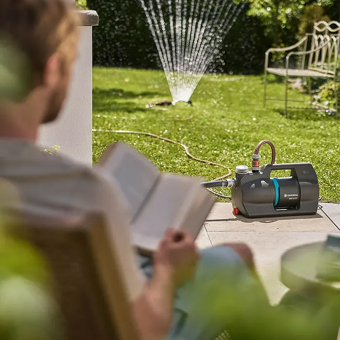 Gardena Hauswasserautomat 4600 Silent - Max. Fördermenge: 4.600 l/h, Max. Druck: 4 bar - Gartenpumpe von Gardena mit Bewässerung im Garten, während eine Person im Hintergrund liest.