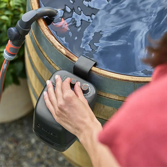 Gardena Power for All 18V Akku-Regenfasspumpe 2500 ComfortAutomatic - Ohne Akku, Max. Fördermenge: 2.400 l/h - Die Gardena-Bewässerungsuhr wird an einem Wasserfass befestigt und von einer Person bedient, um die Gartenbewässerung zu steuern.