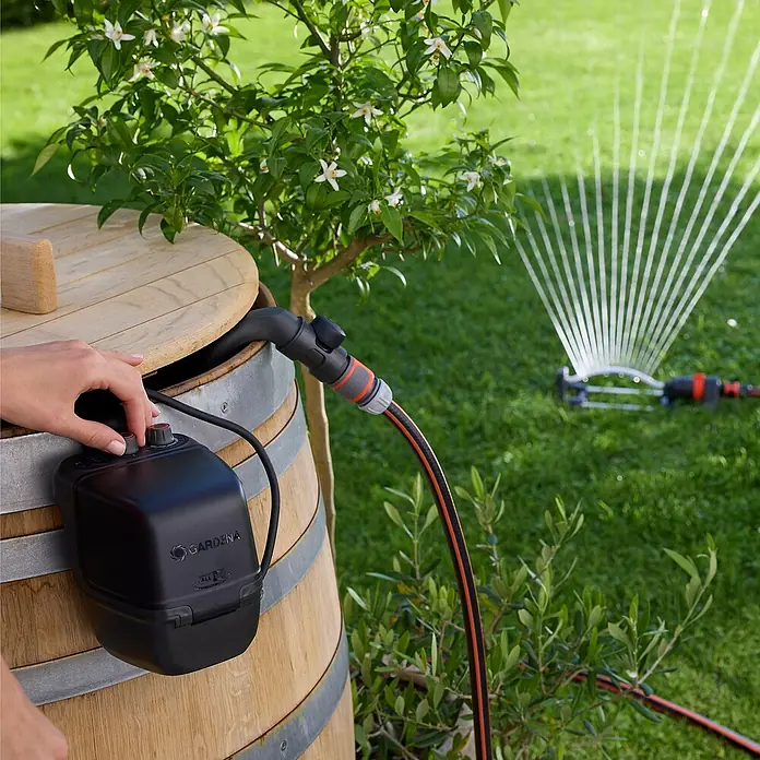 Gardena Power for All 18V Akku-Regenfasspumpe 2600 Comfort - Ohne Akku, Max. Fördermenge: 2.600 l/h - Gardena Regenfasspumpe, die an ein Regenfass angeschlossen ist und ein Rasensprenger auf einem grünen Rasen bewässert.