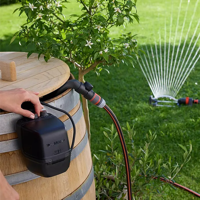Gardena Power for All 18V Akku-Regenfasspumpe 2500 ComfortAutomatic - Ohne Akku, Max. Fördermenge: 2.400 l/h - Das Bild zeigt eine Gardena Regenfasspumpe, die an ein Holzfass angeschlossen ist und einen Rasensprenger speist. Bewässerung leicht gemacht!