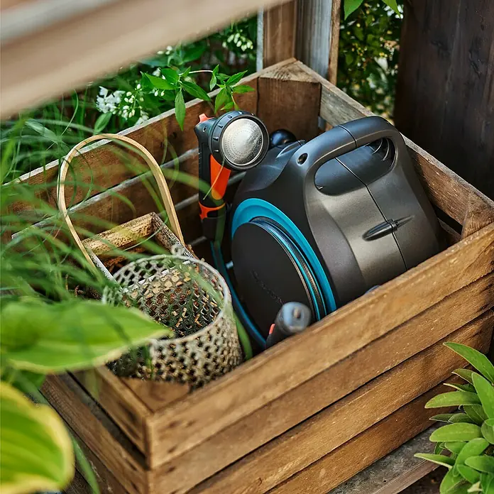 Gardena Schlauchbox MoveMatic - Schlauchlänge: 12,5 m - Eine hölzerne Kiste mit einer Gardena Schlauchbox, einer Gießkanne und anderem Gartenzubehör, umgeben von üppigem Grün.