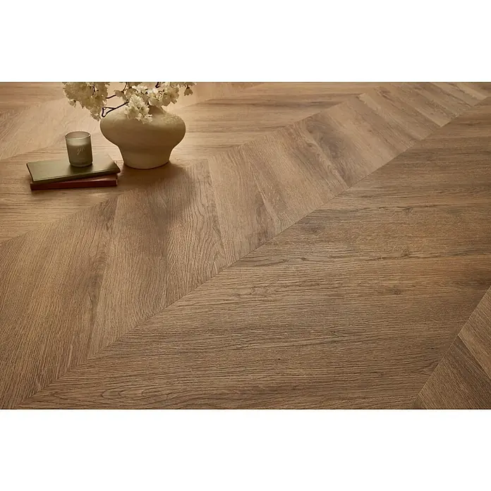 Vinylboden SPC Chevron - Garda Eiche, 120 x 600 x 4,5 mm - Detailaufnahme eines Chevron-Holzfußbodens, der ein elegantes und modernes Design mit warmen Farbtönen zeigt. Eine Vase mit Blumen und Bücher zieren den Boden.