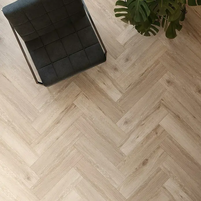 Vinylboden SPC Herringbone - Tucson Eiche, Fischgrät, 148 x 592 x 4,5 mm - Ansicht von oben auf einen grauen Sessel und eine grüne Pflanze auf einem hellen Fischgrätparkettboden, der ein modernes Wohnambiente ausstrahlt.