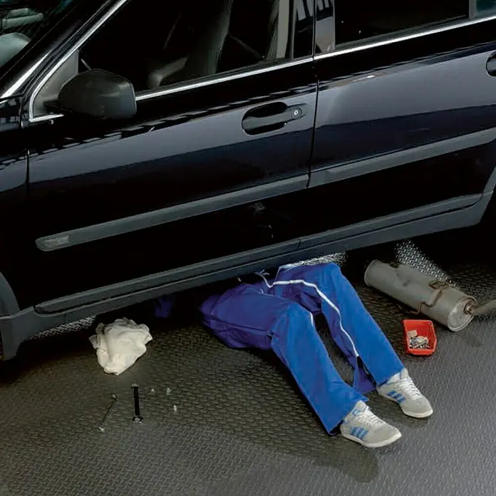 Mottez Universalmatte - Grau, 60 x 60 cm - Eine Person, die unter einem schwarzen Auto in blauer Arbeitskleidung liegt und Reparaturen an einer Auspuffanlage durchführt.