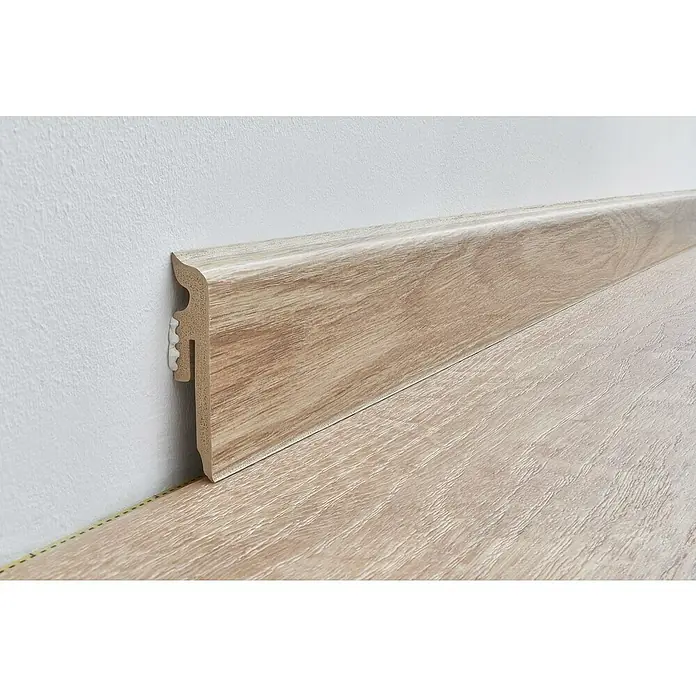 Sockelleiste Vigo - 220 x 1,5 x 6 cm, Sherman Eiche - Helle Holzfußleiste an einer weißen Wand, die den Boden ziert. Die Fußleiste bietet Schutz und ein ästhetisches Erscheinungsbild.