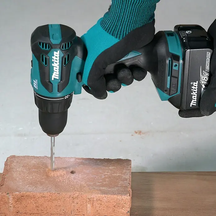 Makita Taladro atornillador percutor de batería DHP490 - 18 V, 2 baterías, 3 Ah, 65 Nm - Un taladro inalámbrico Makita perfora un ladrillo, sujetado por manos enguantadas. La herramienta eléctrica es potente y eficiente.