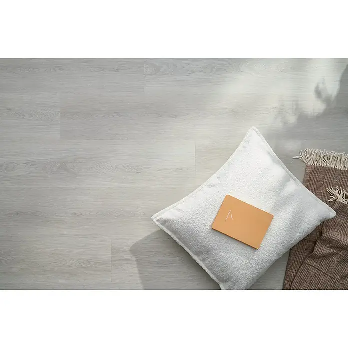 Vinylboden SPC Silent - Takeda Eiche, Landhausdiele, 1.220 x 229 x 5,3 mm - Ein cremefarbenes Kissen mit einem Notizbuch darauf, ergänzt durch eine braune Decke, alles auf einem hellen Holzboden arrangiert.
