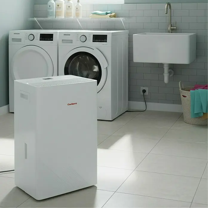 Corberó Deshumidificador eléctrico CDG20 - 380 W, Potencia de deshumidificación: 20 l/día, L x An x Al: 23,5 x 31 x 50 cm, Blanco - Imagen de un deshumidificador Cerbero en un cuarto de lavado cerca de una lavadora y secadora. La unidad es compacta y blanca.