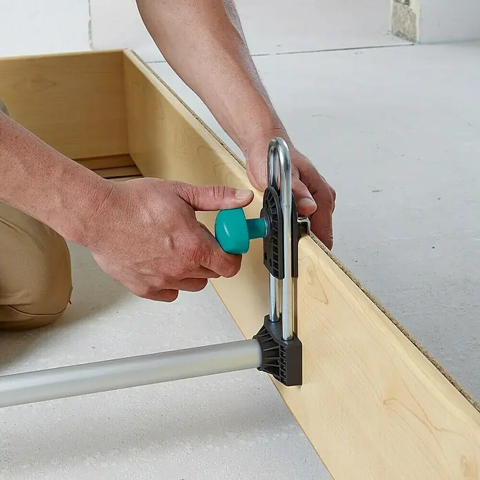Wolfcraft Soporte para marcos de puerta Easy - Específico para: Puertas de 600 - 1.000 mm de ancho - Una persona está sujetando una tabla de madera a un marco con una abrazadera de una mano de Wolfcraft. La herramienta es azul y plateada.
