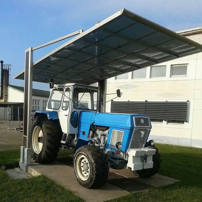 Ximax Carport Miro Next Typ 60 - Außenmaß inkl. Dachüberstand (B x T): 3,03 x 5,79 m, Edelstahl-Look - Ein blauer und weißer Zetor-Traktor, geparkt unter einer Überdachung. Er steht auf einer Betonfläche, umgeben von grünem Gras.
