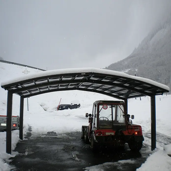 Ximax Carport Portoforte Typ 80 - M Ausführung - Außenmaß inkl. Dachüberstand (B x T): 6,03 x 5,56 m, Mattbraun - Carport im Winter mit Schnee bedeckt, darunter ein roter Traktor. Im Hintergrund ein eingeschneites Auto.