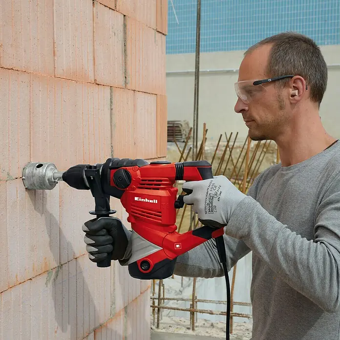Einhell Kombihammer TC-RH 1600 - 1.600 W, 4 J, SDS-Plus-Aufnahme - Ein Mann bohrt mit einer Einhell-Bohrkrone ein Loch in eine Ziegelwand, wobei er Schutzbrille und Handschuhe trägt.