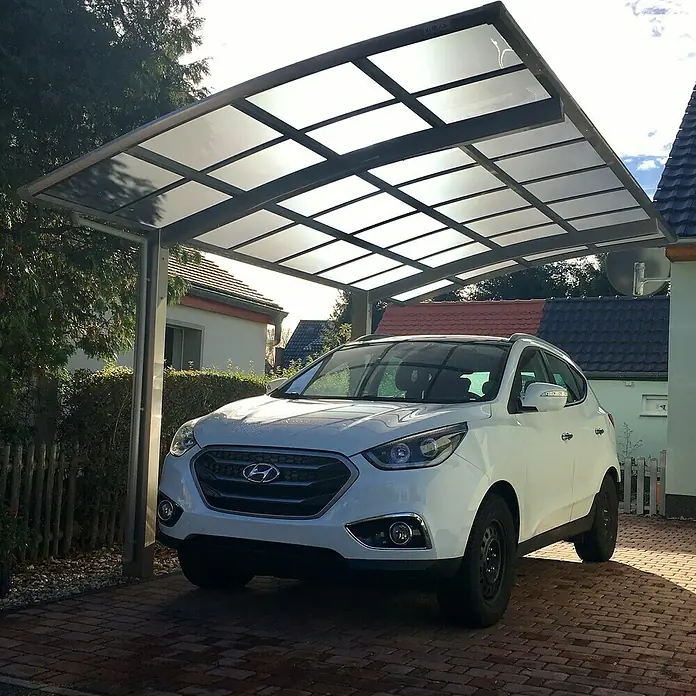 Ximax Carport PF60 - Außenmaß inkl. Dachüberstand (B x T): 3,01 x 5,56 m, Einzelcarport, Edelstahl-Look - Das Bild zeigt ein weißes Auto, das unter einem modernen Carport mit transparentem Dach geparkt ist, das Schutz vor Witterungseinflüssen bietet.