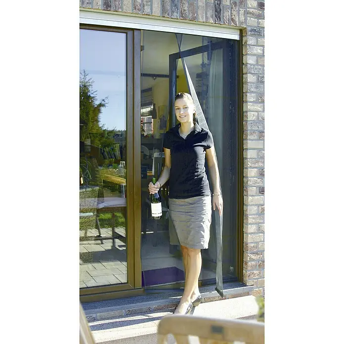 Schellenberg Cortina con protección contra insectos - An x Al: 70 x 220 cm, Montaje de la mosquitera: Pegado - Una mujer entra en una casa con una botella de vino a través de una puerta mosquitera magnética, una solución práctica para la protección contra insectos.