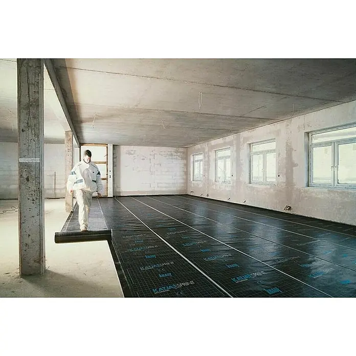 Knauf Katja Sprint Abdichtungsbahn - 32 m x 1,25 m x 0,9 mm - Ein Mann rollt eine schwarze KATIA Sprint Unterlagsbahn mit Rastermuster auf einem unfertigen Betonboden in einem Raum mit Fenstern aus.