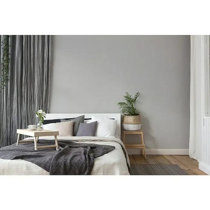 A.S. Création Hygge 3 Vliestapete - Hellgrau II, Uni, 10,05 x 0,53 m - Elegantes Schlafzimmer mit einem Holzbett-Tablett mit Blumen und Getränken auf einer grauen Decke. Pflanze auf Holzschemel neben dem Bett.