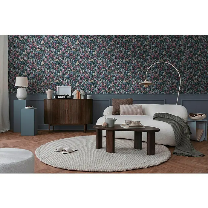 A.S. Création Cosmoliving by Cosmopolitan Vliestapete Blüte - Blau-Grau-Pink, Floral, 10,05 x 0,53 m - Stilvolles Wohnzimmer mit dunkler Blumentapete, einem weißen Sofa und einem runden Teppich. Die Einrichtung ist modern und gemütlich.