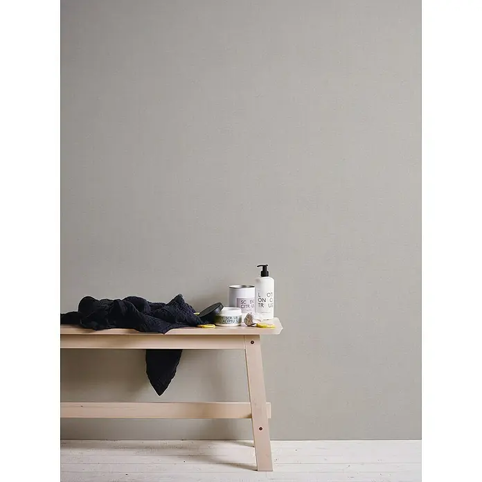 A.S. Création Hygge 3 Vliestapete - Beigegrau, Uni, 10,05 x 0,53 m - Stillleben mit Badutensilien auf einem Hocker vor einer grauen Wand. Kosmetikprodukte und ein dunkles Handtuch sind auf dem Hocker platziert.
