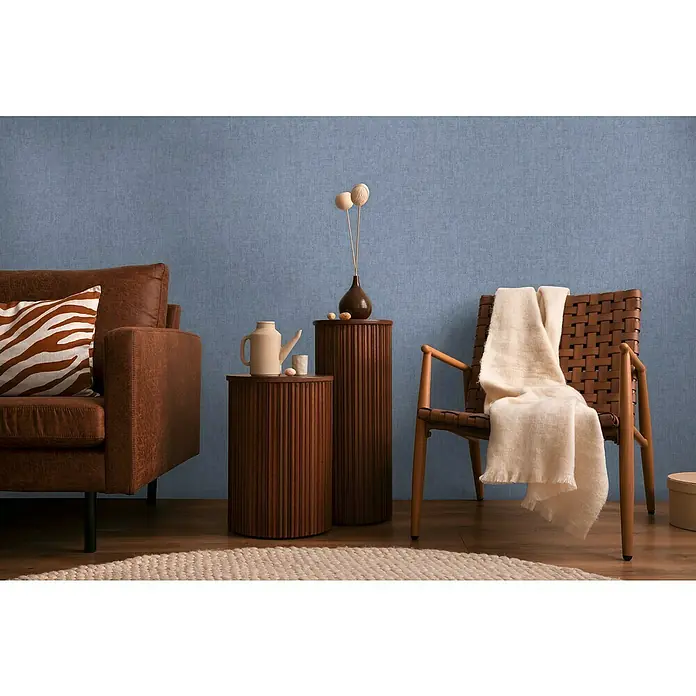 A.S. Création Cosmoliving by Cosmopolitan Vliestapete - Mittelblau, Uni, 10,05 x 0,53 m - Stilvolles Wohnzimmer-Setup mit Ledersofa, Holztisch und Webstuhl-Stuhl, ergänzt durch eine dekorative Vase und eine Decke.