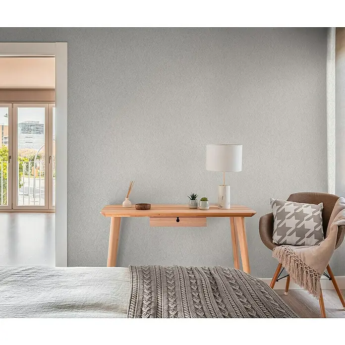 A.S. Création Hygge 3 Vliestapete - Hellgrau, Uni, 10,05 x 0,53 m - Nahaufnahme eines Schlafzimmers mit einem Holztisch, einer Lampe und einem Stuhl mit Kissen, der eine gemütliche und stilvolle Atmosphäre ausstrahlt.