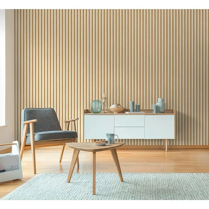 A.S. Création Hygge 3 Vliestapete Paneele - Creme-Braun, Holzoptik, 10,05 x 0,53 m - Stilvolles Wohnzimmer mit hellem Holzvertäfelung. Moderner Sessel, Sideboard und ein kleiner Tisch mit Tasse und Teller schmücken den Raum.