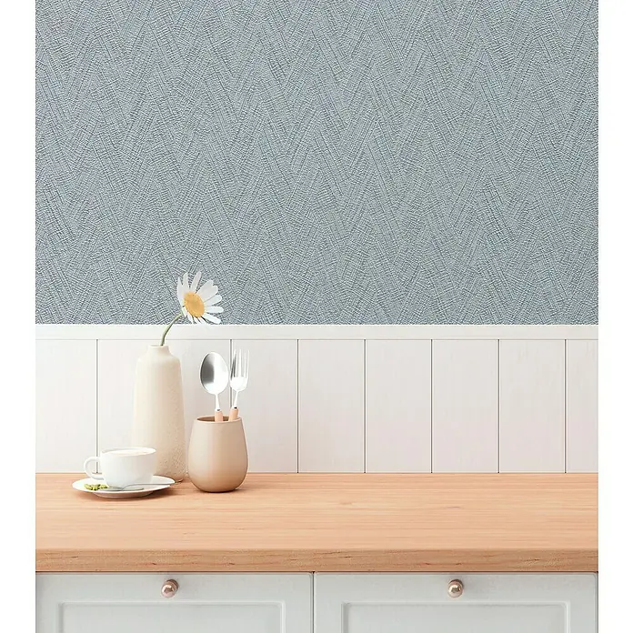 A.S. Création Hygge 3 Vliestapete - Grau-Blau, Uni, 10,05 x 0,53 m - Nahaufnahme eines hellen Küchentisches mit Geschirr, einer Tasse und einer Vase, vor einer strukturierten blauen Wand.
