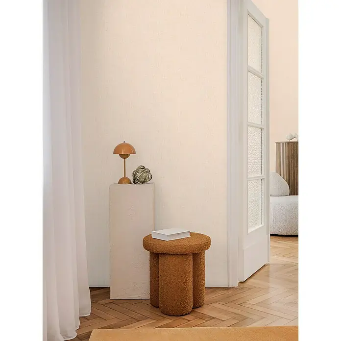 A.S. Création Cosmoliving by Cosmopolitan Vliestapete - Mittelbeige, Uni, 10,05 x 0,53 m - Einladendes Interieur mit einem runden, braunen Hocker mit drei Beinen und einem Buch, das eine gemütliche und stilvolle Atmosphäre schafft.