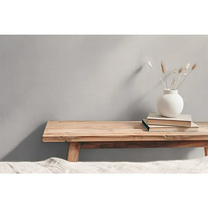 A.S. Création Hygge 3 Vliestapete - Hellgrau I, Uni, 10,05 x 0,53 m - Schlichte Inneneinrichtung mit einer weißen Vase und Büchern auf einer Holzbank vor einer grauen Wand.