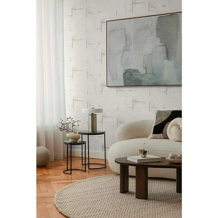 A.S. Création Cosmoliving by Cosmopolitan Vliestapete Beton - Grau-Weiß, Betonoptik, 10,05 x 0,53 m - Wohnzimmer mit einem runden Teppich, einem cremefarbenen Sofa und einem Couchtisch aus dunklem Holz. Abstraktes Gemälde und eine moderne Lampe.