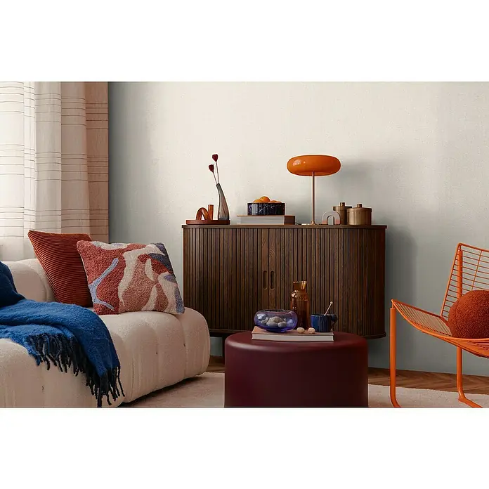 A.S. Création Cosmoliving by Cosmopolitan Vliestapete - Cremebeige, Uni, 10,05 x 0,53 m - Wohnzimmer-Szene mit einem Holzschrank, einem orangefarbenen Sessel und einem bequemen Sofa mit Kissen und Decke.